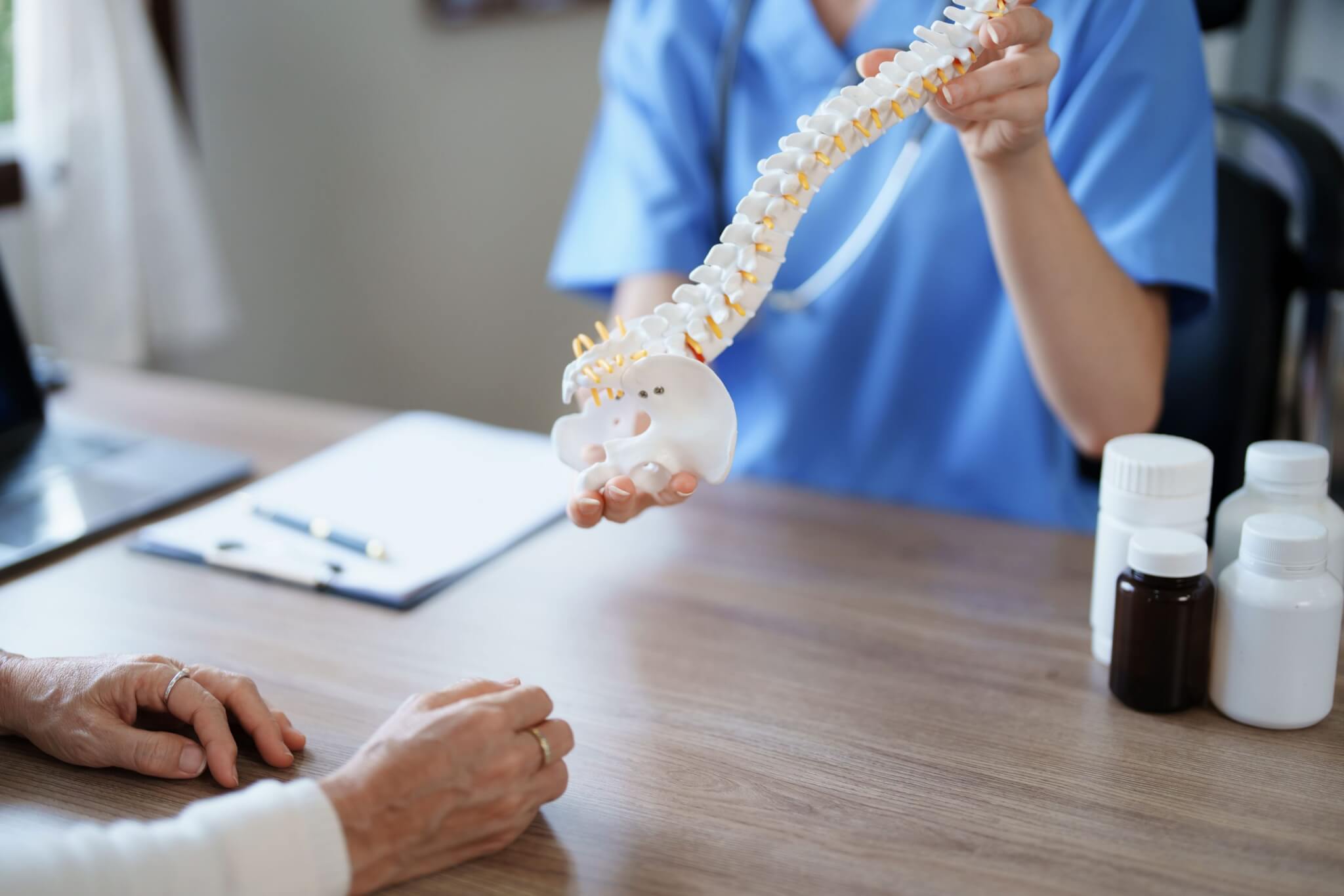 Clinician in blue scrubs displays a curved spine and pelvis model across a desk
